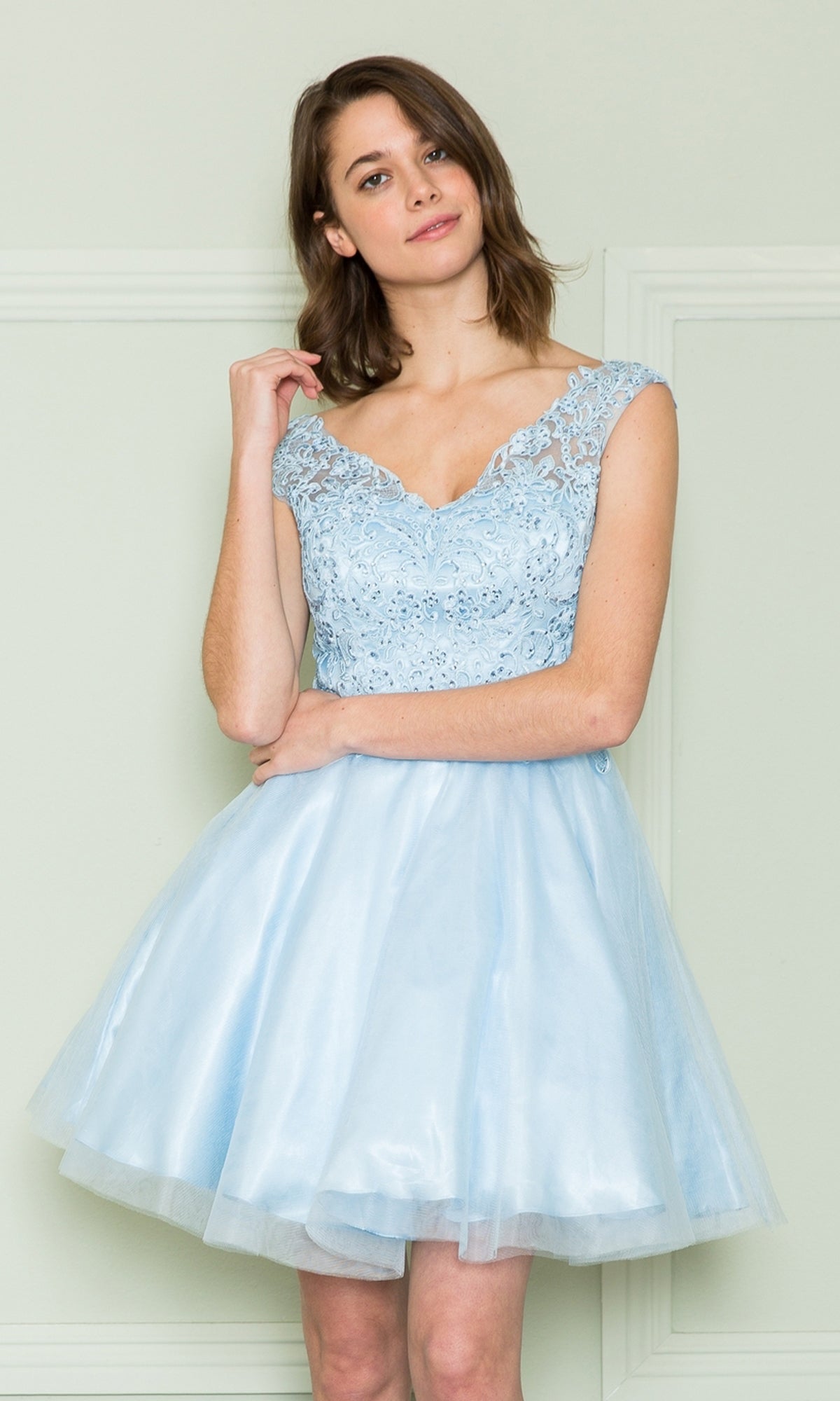 A woman wearing the Lace-Bodice Short Babydoll Homecoming Dress 8902, featuring a sleeveless light blue lace corset bodice and tulle skirt, stands against a pale wall and smiles softly at the camera.